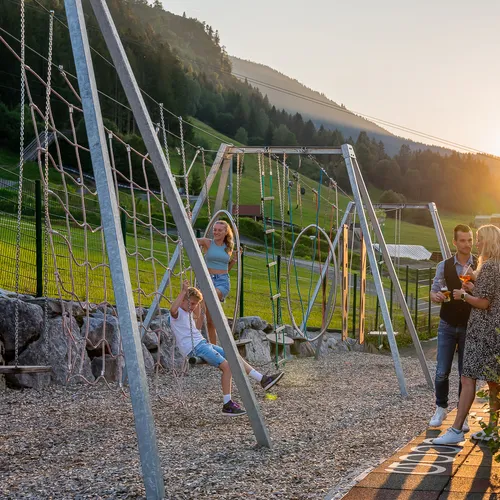 Familie auf Spielplatz in den Bergen bei Sonnenuntergang mit Schaukeln und Getränken